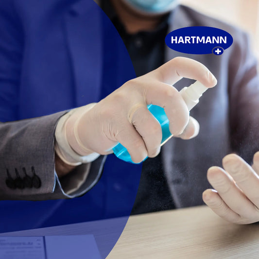 Eine Person in einem Anzug, mit Handschuhen und einer Gesichtsmaske sprüht eine blaue Flüssigkeit aus einer Flasche auf ihre Hand. Das HARTMANN-Logo erscheint in der oberen rechten Ecke des Bildes.