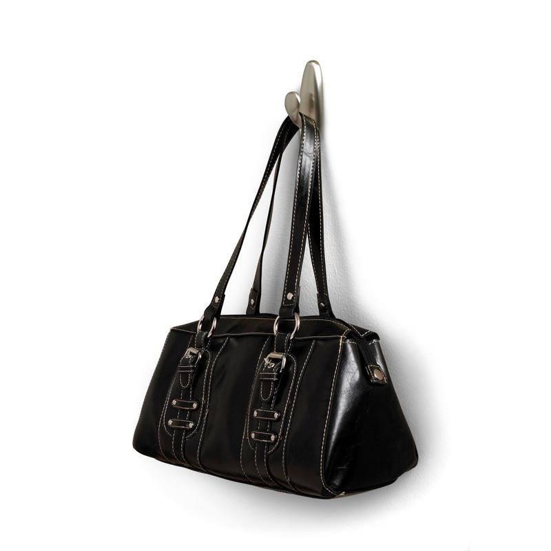 De un cârlig metalic Command™ „clasic” de la 3M Deutschland GmbH este agățată o geantă de mână din piele neagră cu mânere duble și catarame decorative. Geanta are cusături albe și o suprafață ușor lucioasă.