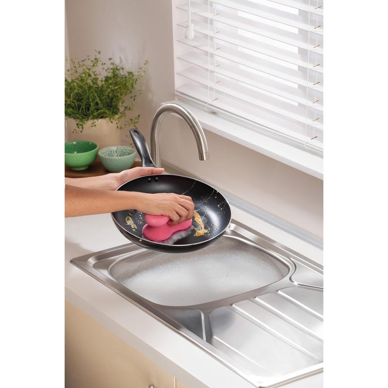 O persoană curăță o tigaie sub apă curgătoare într-o bucătărie modernă cu un burete de curățare Scotch-Brite® Delicate Color Comfort care nu zgârie (2 bucăți) de la 3M Deutschland GmbH - ideal pentru suprafețe delicate. Boluri verzi și o plantă sunt așezate pe blat.