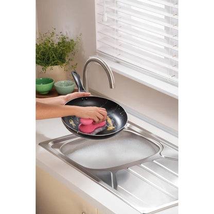 O persoană curăță o tigaie sub apă curgătoare într-o bucătărie modernă cu un burete de curățare Scotch-Brite® Delicate Color Comfort care nu zgârie (2 bucăți) de la 3M Deutschland GmbH - ideal pentru suprafețe delicate. Boluri verzi și o plantă sunt așezate pe blat.