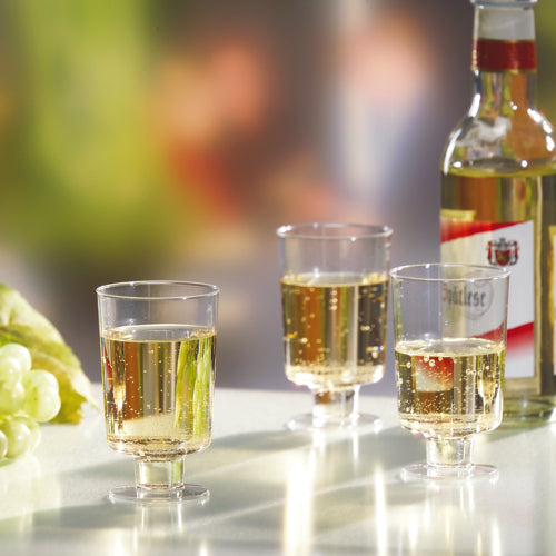 Trei pahare Starpak cu picior pentru vin alb (PS, 0,1l, clar ca sticla) sunt umplute și stau lângă o sticlă de vin alb și struguri verzi pe o suprafață albă în fața unui fundal neclar.