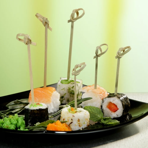 Este prezentată o farfurie neagră cu diverse tipuri de sushi, fiecare înfipt cu un PAPSTAR 250 Fingerfood-Picker „pure“ de la PAPSTAR GmbH. Totul este decorat cu icre, frunze de legume și wasabi; fundalul este în tonuri blânde de verde și galben.