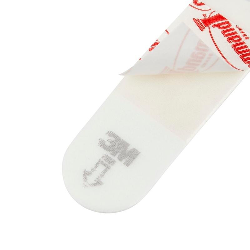 Eine Nahaufnahme eines Kleine Command™ Strips S, 17022N von 3M Deutschland GmbH zeigt seine rechteckige Form, wobei ein Ende des Schutzpapiers abgezogen ist und der Klebstoff zum Vorschein kommt. "MC" und ein Pfeil sind aufgedruckt, um eine einfache und beschädigungsfreie Verwendung an der Wand zu ermöglichen.