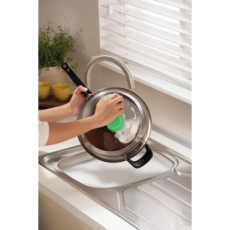 Cineva curăță un capac de vas din metal deasupra unei chiuvete cu spumă folosind buretele de curățare Scotch-Brite® Classic de la 3M Deutschland GmbH, lângă o fereastră cu jaluzele albe și un tejghea cu o plantă și boluri galbene stivuite.