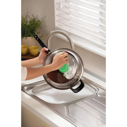 Cineva curăță un capac de vas din metal deasupra unei chiuvete cu spumă folosind buretele de curățare Scotch-Brite® Classic de la 3M Deutschland GmbH, lângă o fereastră cu jaluzele albe și un tejghea cu o plantă și boluri galbene stivuite.