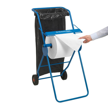 Un suport metalic albastru ține un șervețel de hârtie WypAll® L10 pentru suprafețe - rolă mare (38 x 23,5 cm) de la Kimberly-Clark GmbH și un sac de gunoi negru; o persoană trage o foaie pentru curățarea suprafețelor.