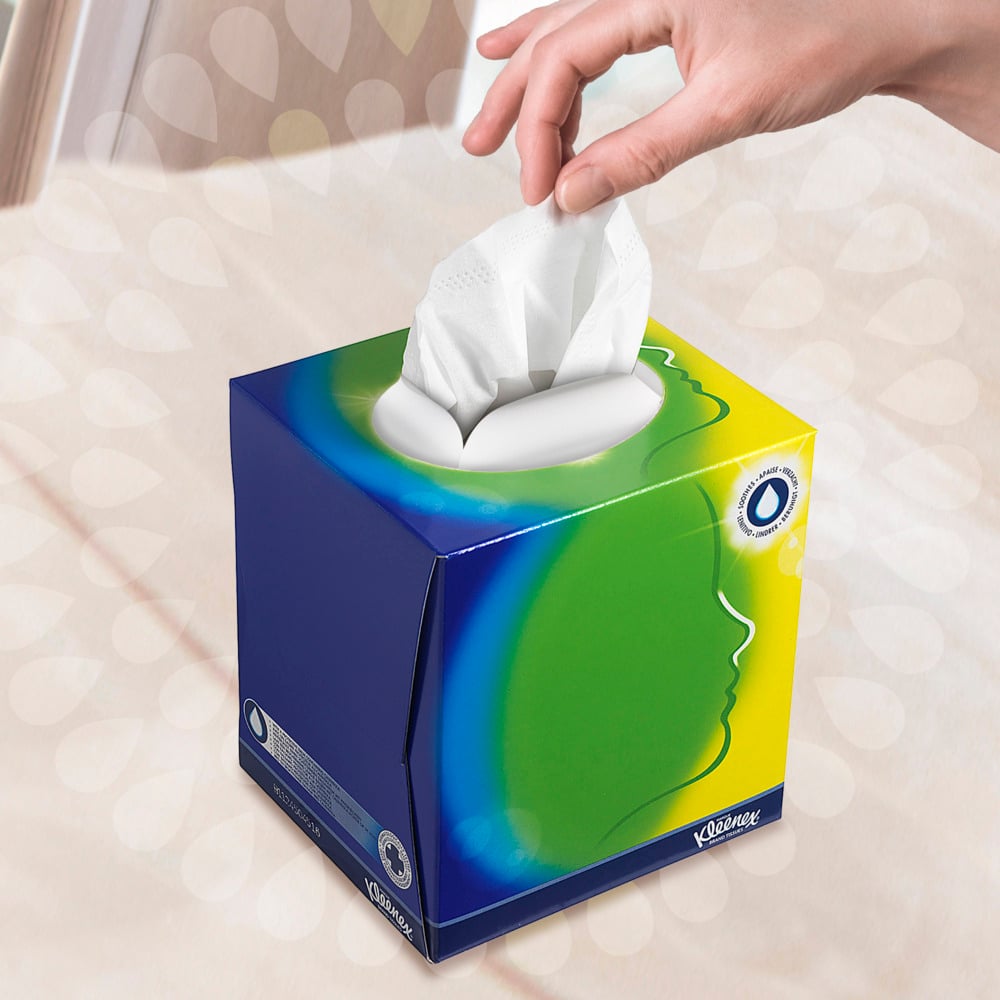 Eine Hand zieht ein Taschentuch aus der Kleenex® Kosmetiktücher - Würfel-Box / Weiß der Kimberly-Clark GmbH mit blauem, grünem und gelbem Design und einer weißen ovalen Öffnung. Ein stilisiertes Profil deutet auf Gesichtspflege vor einem sanft verschwommenen Hintergrund hin.