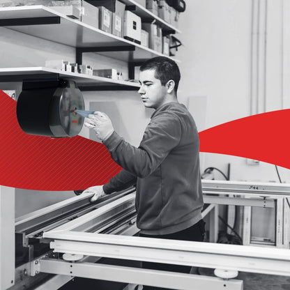 Într-un atelier, o persoană folosește un dispozitiv montat pe perete în apropierea unor rafturi cu șervețele de hârtie WypAll® L10 pentru alimente și igienă Centrefeed (6 role) de la Kimberly-Clark GmbH; deasupra se află cutii și un swoosh roșu accentuează fundalul.