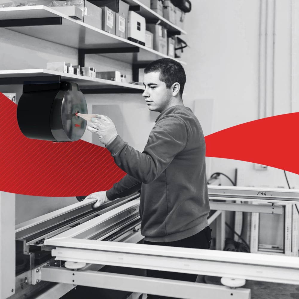 Într-un atelier, o persoană extrage un șervețel de hârtie WypAll® L20 de la Kimberly-Clark GmbH prin extragere centrală din distribuitorul de perete și curăță suprafețe de lucru industriale; un element grafic roșu traversează imaginea pe mijloc.