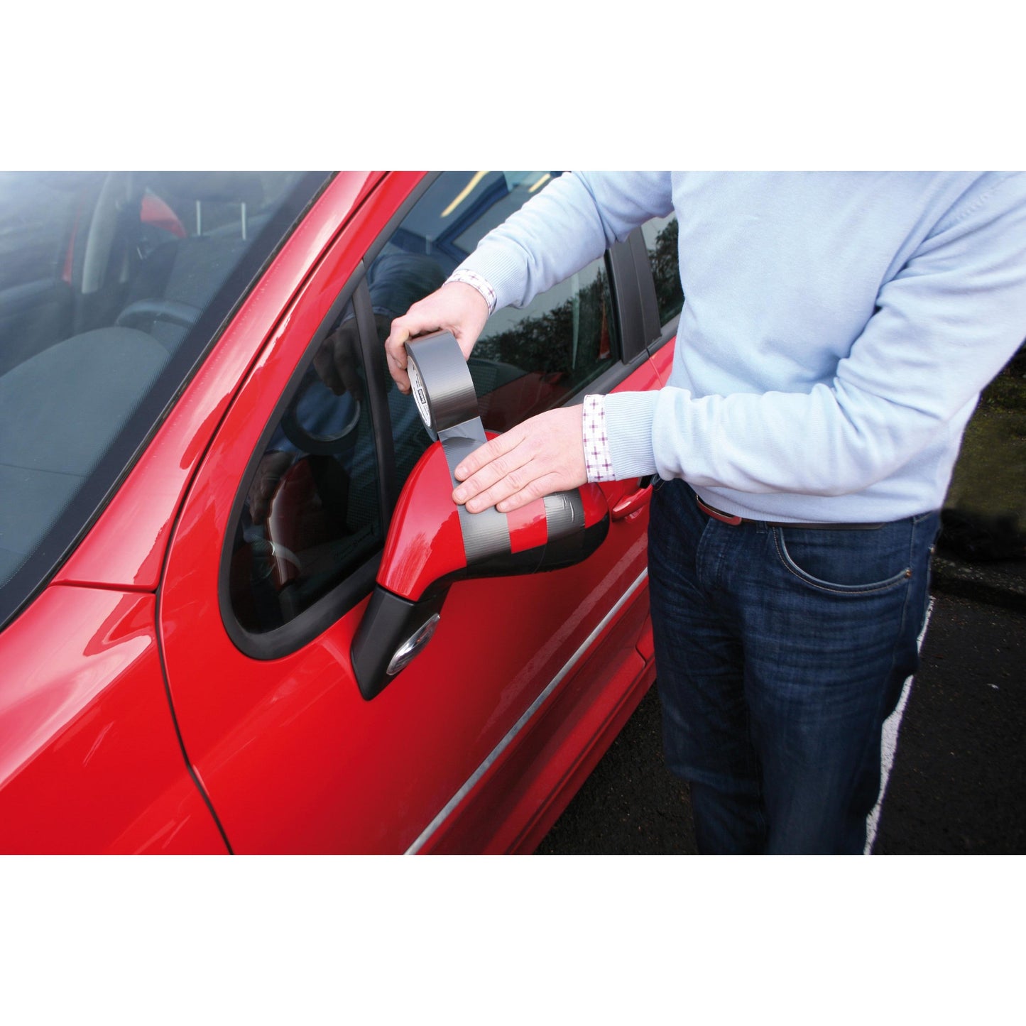 Eine Person in einem hellblauen Pullover verwendet das Scotch® Extremium™ No Residue Hochleistungs-Gewebeklebeband (18,2 m x 48 mm) von 3M, um den beschädigten Seitenspiegel eines roten Autos zu befestigen, während sie an der Fahrertür steht. Das graue Band ist effektiv und hinterlässt keine Rückstände.