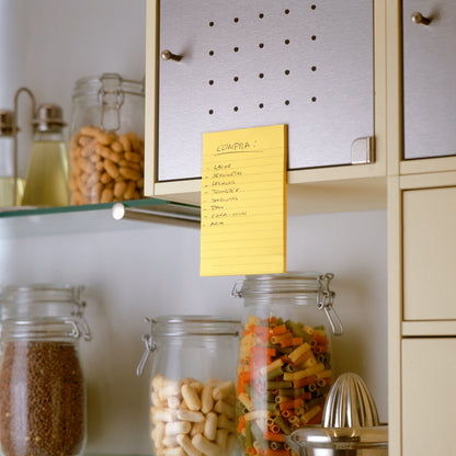 O listă de cumpărături scrisă de mână în limba spaniolă se află pe un dulap de bucătărie bej folosind o notiță Post-it® Super Sticky în format mare de la 3M Deutschland GmbH, Ultra Galben (101 x 152 mm), peste borcane cu paste, fasole și cereale. Pe rafturile adiacente se află alte borcane și ustensile de bucătărie.