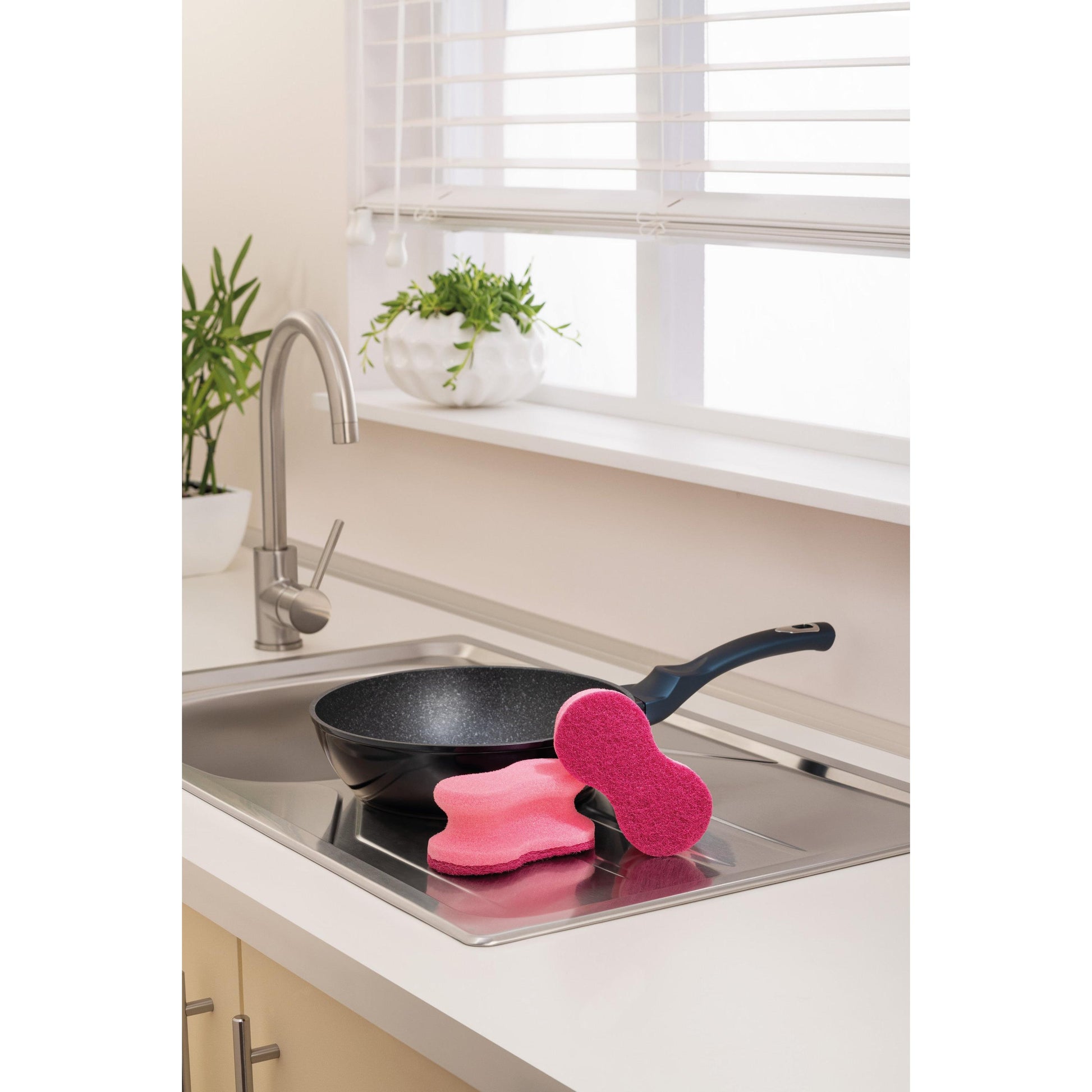 Eine schwarze Bratpfanne liegt auf einer Küchenspüle neben zwei rosafarbenen Scotch-Brite® Delicate Color Comfort kratzfreien Schwämmen (2 pro Packung) der 3M Deutschland GmbH. Im Hintergrund befinden sich Topfpflanzen und ein Fenster mit weißen Jalousien.