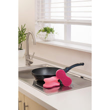 Eine schwarze Bratpfanne liegt in einer Küchenspüle, auf deren Rand zwei rosafarbene Scotch-Brite® Delicate Color Comfort nicht kratzender Reinigungsschwamm (2 Stück, 3M Deutschland GmbH) liegen; die weiße Arbeitsplatte und Topfpflanzen in der Nähe vervollständigen die Szene.