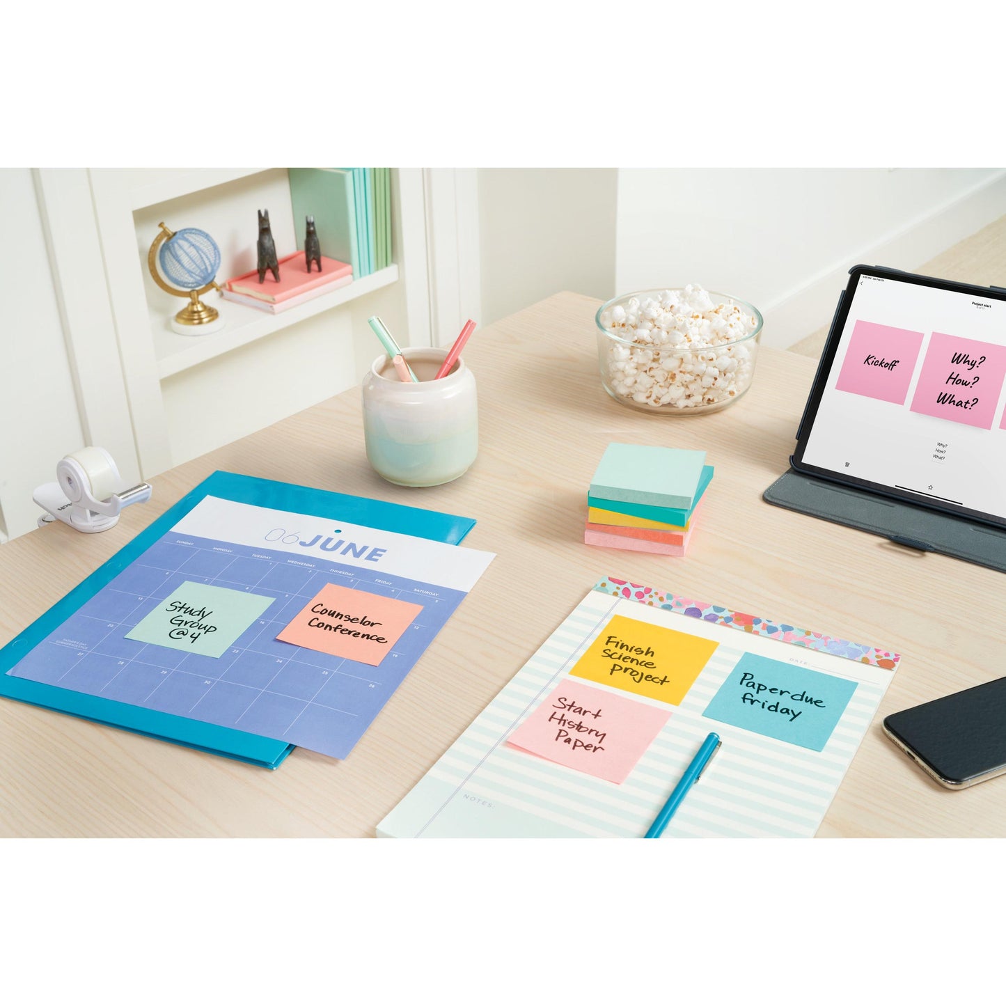 Un birou cu un calendar de iunie, creioane pastelate într-un pahar, 3M Post-it® Notes Beachside Collection (38x51mm, 100 foi/bloc, 12 blocuri/pachet, ambalaj de carton, 100% PEFC), un distribuitor de bandă adezivă, o tablă deschisă cu note - și un mic glob pe raft.