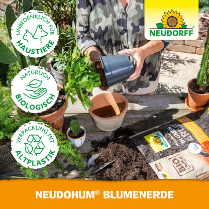 Eine Person beim Umtopfen einer Pflanze an einem Tisch mit Erde, getopften Gartenpflanzen, Gartengeräten, Blumenerde-Taschen und einer NeudoHum BlumenErde-Tasche von Neudorff Shop. Neben dem Neudorff-Logo erscheinen die Symbole für Umweltfreundlichkeit und Haustierfreundlichkeit.