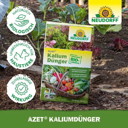 Eine Schachtel Azet KaliumDünger aus dem Neudorff Shop steht auf der Gartenerde zwischen Gemüse und Grünpflanzen und zeigt Produktinfos und Zertifizierungen. Ideal zum Düngen von Pflanzen und zur Vorbeugung von Kaliummangel.