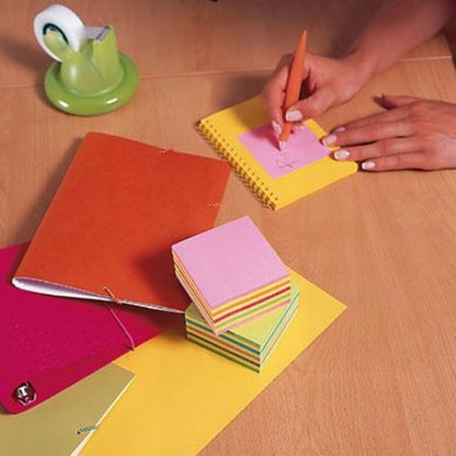 La un birou de lemn, cineva scrie cu un stilou portocaliu pe un cub de notițe Post-it® roz (76 mm x 76 mm, 450 foi/bloc, 100% PEFC) de la 3M Deutschland GmbH. În apropiere se află un caiet roșu, un suport de bandă adezivă verde, un clopoțel argintiu și alte foi colorate.