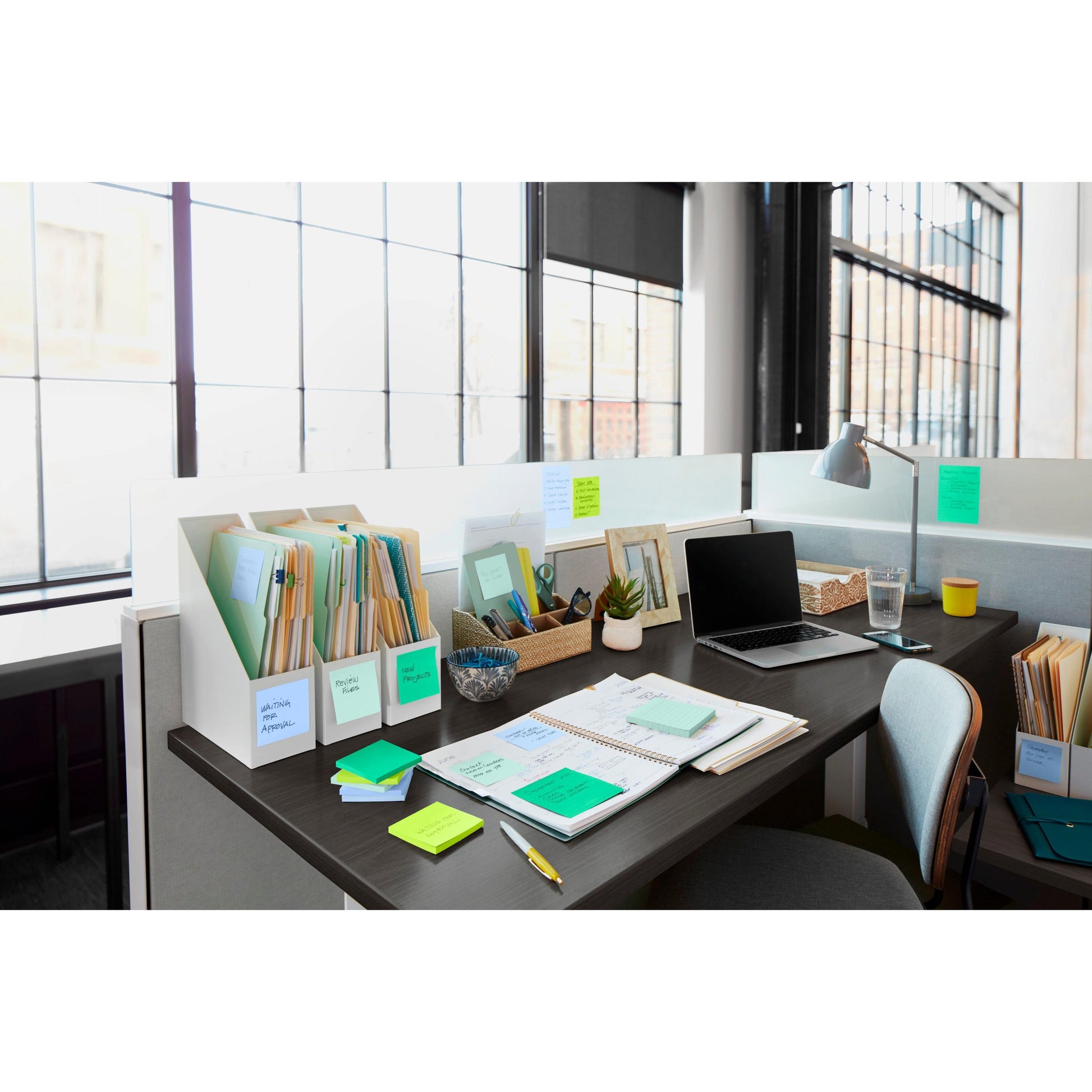 Ein moderner Bürotisch mit Post-it® Super Sticky Notes (76 mm x 76 mm, 90 Blatt/Block, 100% PEFC) der 3M Deutschland GmbH, Ordnern, Notizbüchern, einem Laptop, einer Lampe, einem Stifthalter, einer kleinen Pflanze und einem Glas Wasser vor sonnenbeschienenen Fenstern.