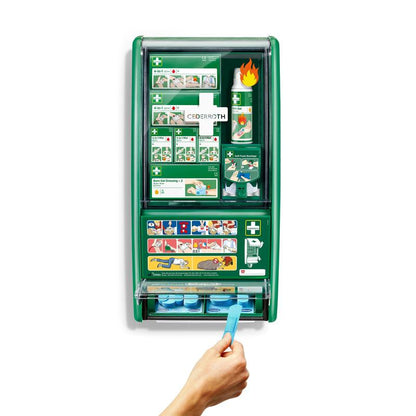 O stație verde de prim ajutor și arsuri Cederroth de la Orkla Wound Care AB este montată pe un perete alb, echipată cu bandaje, gel pentru arsuri, soluție pentru clătirea ochilor și altele. O mână trage un bandaj adeziv albastru din distribuitorul inferior al cutiei de 1 buc.