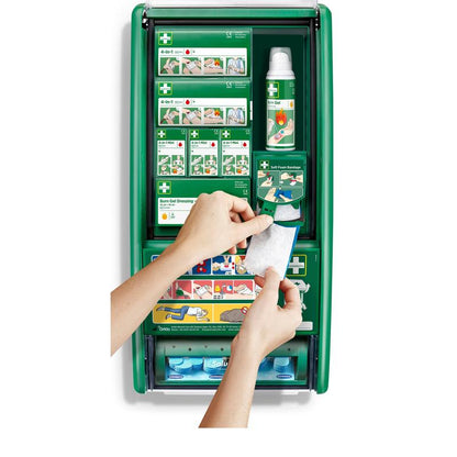 O persoană ține un bandaj cu gel pentru arsuri în fața stației de prim ajutor și arsuri Cederroth (1 pachet) de la Orkla Wound Care AB, o stație verde de prim ajutor cu materiale pentru arsuri, bandaje, sticle și instrucțiuni.