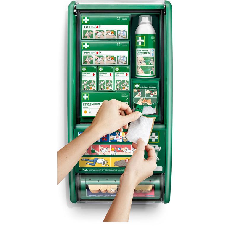 Eine Person hält einen Verband vor einer Cederroth Erste-Hilfe-Station von Orkla Wound Care AB, die mit Verbänden, Verbrennungsgel, Kochsalzlösung und Binden bestückt ist. Illustrierte Anweisungen sind auf den Fächern im Inneren zu sehen.