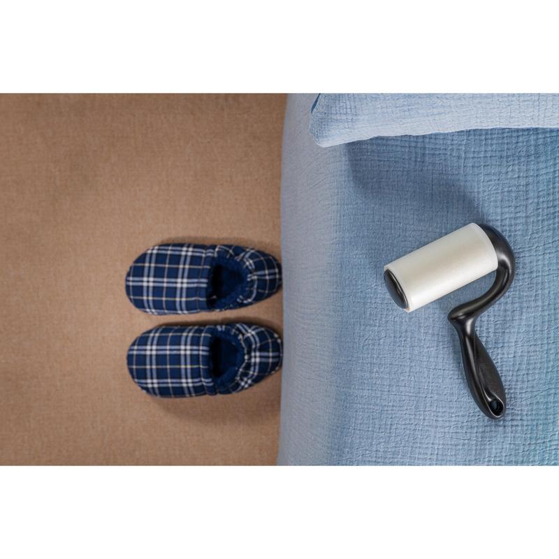 O pereche de papuci de casă în carouri albastre stă pe un covor bej lângă un pat cu o cuvertură albastru deschis, în timp ce roleul de scame Scotch-Brite™ Everyday Clean pentru suprafețe plane (56 de foi, 1/pachet) de la 3M Deutschland GmbH se află pe pat și este gata de utilizare.