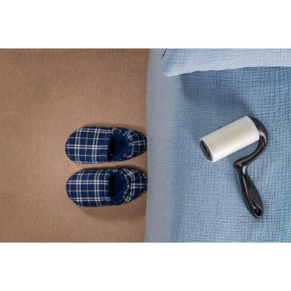 O pereche de papuci de casă în carouri albastre stă pe un covor bej lângă un pat cu o cuvertură albastru deschis, în timp ce roleul de scame Scotch-Brite™ Everyday Clean pentru suprafețe plane (56 de foi, 1/pachet) de la 3M Deutschland GmbH se află pe pat și este gata de utilizare.