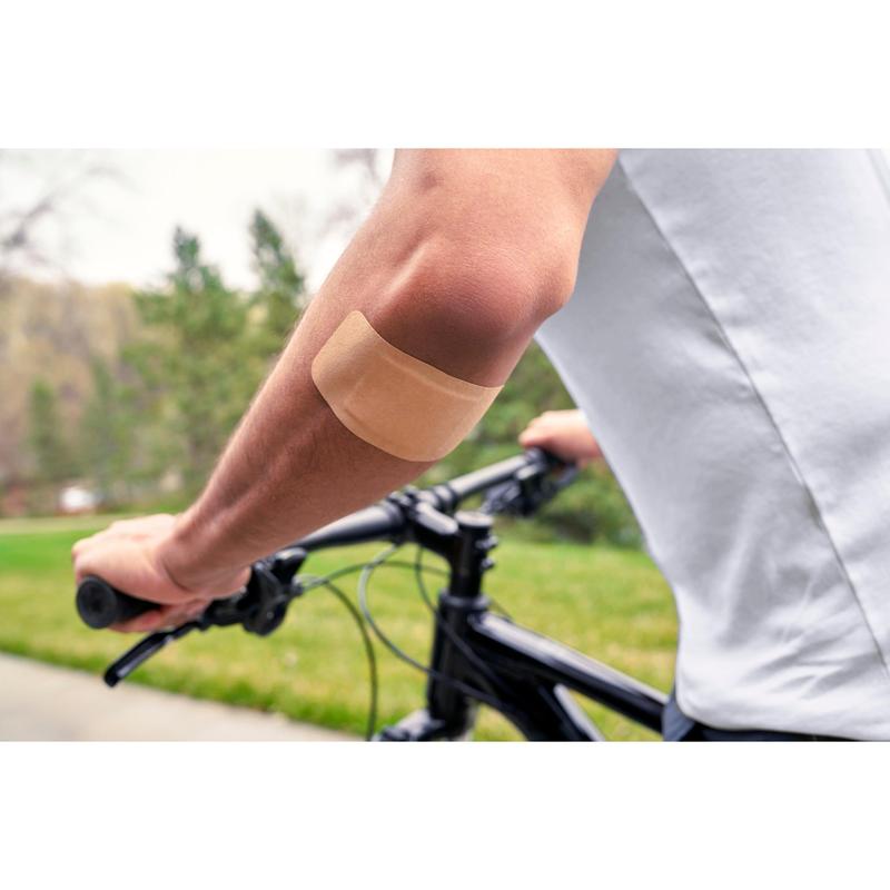 O persoană care poartă o cămașă albă și merge pe bicicletă în aer liber are un plasture Nexcare™ Ultra Stretch MAXI Comfort Flexible (50 mm x 101 mm) de la 3M Deutschland GmbH pe brațul gol; în fundal se văd iarbă verde și copaci.
