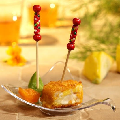 Pe un platou transparent, în formă de frunză, se află un cub de mâncare prăjită și pane cu un PAPSTAR 250 de scobitori Fingerfood ale PAPSTAR GmbH, alături de o felie de portocală și o felie verde. În fața unui fundal ușor neclar se pot vedea o felie de lămâie și verdețuri.