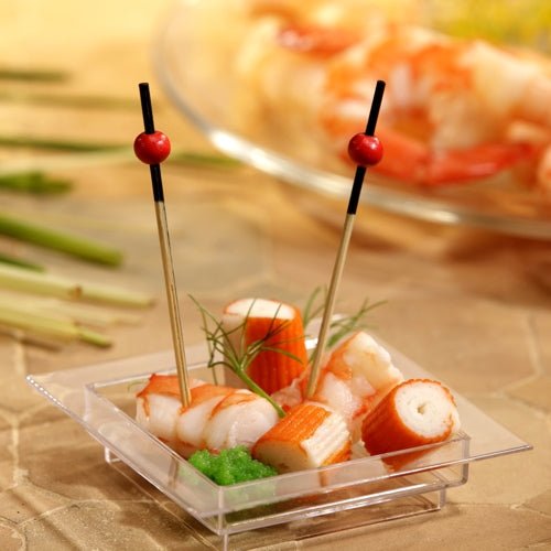Pe un platou transparent, pătrat, se află imitație de crab, creveți, o garnitură verde și două PAPSTAR 250 de scobitori Fingerfood ale PAPSTAR GmbH cu mărgele roșii. În fundal, se pot vedea creveți neclar pe o farfurie.