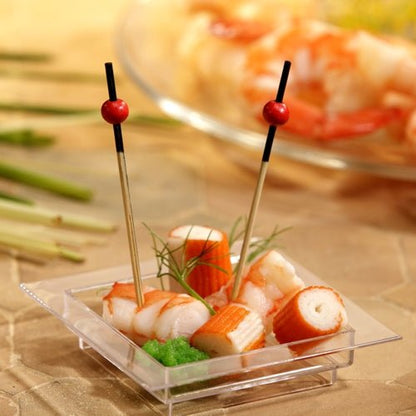 Pe un platou transparent, pătrat, se află imitație de crab, creveți, o garnitură verde și două PAPSTAR 250 de scobitori Fingerfood ale PAPSTAR GmbH cu mărgele roșii. În fundal, se pot vedea creveți neclar pe o farfurie.