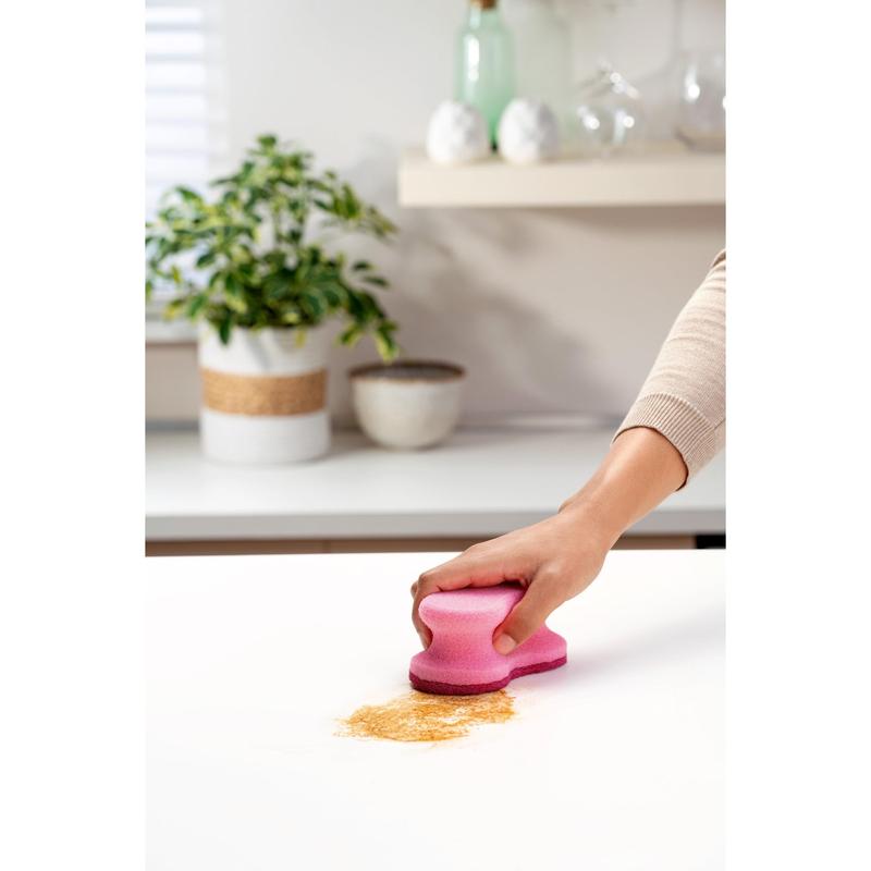 O mână folosește un burete de curățare roz Scotch-Brite® Delicate Color Comfort care nu zgârie (2 bucăți, 3M Deutschland GmbH) pentru a curăța pulbere maro de pe un blat alb - perfect pentru suprafețe delicate. O plantă și boluri în fundal.