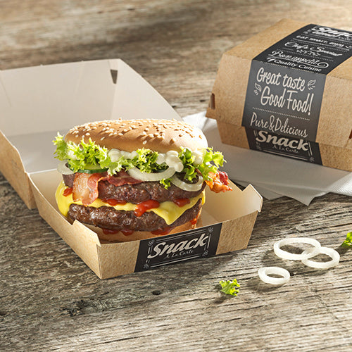 Un cheeseburger cu salată, roșie, bacon, ceapă și sos se află într-o PAPSTAR 50 de cutii de burger din carton "pure" de la PAPSTAR GmbH pe o masă de lemn. Alături, o cutie închisă cu inscripția "Good Food" și inele de ceapă.