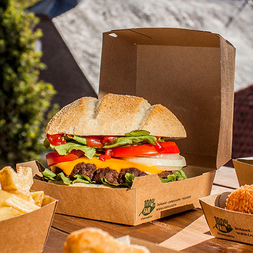 Un cheeseburger cu salată, roșii, ceapă și brânză Cheddar este așezat în aer liber într-o cutie de burger din carton PAPSTAR GmbH (9x18,5x18,5 cm, "100% Fair", 25 bucăți). Cartofii prăjiți și alte garnituri sunt servite separat lângă.