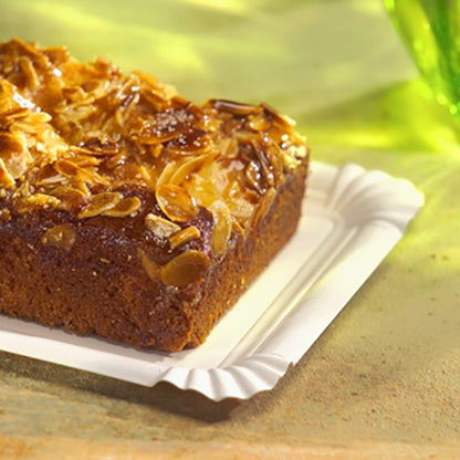 Pe tava de carton PAPSTAR dreptunghiulară 13x20 cm, albă, "pure" pentru petreceri (12 bucăți) de la PAPSTAR GmbH se află un tort dreptunghiular cu migdale feliate - o opțiune de servire ecologică, elegantă, pe o suprafață deschisă cu un pahar verde în spate.