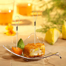 Un bol de sticlă prezintă un cub de aperitiv pane, prăjit, cu un PAPSTAR Fingerfood Picker "Loop" (10 cm), un segment de portocală și o garnitură verde. În fundal, felii de lămâie și două băuturi ale PAPSTAR GmbH sunt neclare.