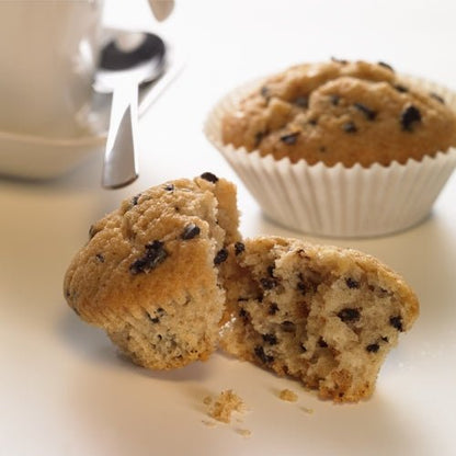Un muffin de ciocolată este tăiat pe jumătate pe o suprafață albă, alături sunt firimituri. În fundal se află un alt muffin întreg într-o PAPSTAR 1000 formă de copt rotundă de la PAPSTAR GmbH, alături de o parte a unei cești de cafea și o lingură.