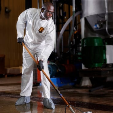 Mit einem KleenGuard® KGA20 Atmungsaktiver Schutzanzug gegen Partikel von Kimberly-Clark GmbH und Handschuhen reinigt eine Person mit einem langstieligen Werkzeug einen nassen Industrieboden, während im Hintergrund Geräte und Rohre zu sehen sind.