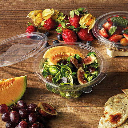 O cutie Starpak 50 pentru delicatese și salate cu capac rabatabil, R-PET transparent rotund, conține o salată de pepene și smochine pe o masă de lemn cu căpșuni, struguri, pâine, cireșe și fructe. Un alt recipient Starpak R-PET cu fructe este în apropiere.