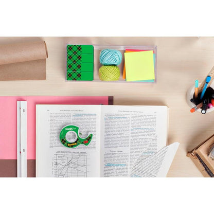 Un spațiu de lucru cu o carte deschisă, hârtie milimetrică, bile de ață colorată, post-it-uri, stilouri într-un pahar, gume de șters, o rolă de hârtie maro și o rolă de bandă invizibilă Scotch® Magic™ de la 3M Deutschland GmbH pe o masă de lemn deschis.