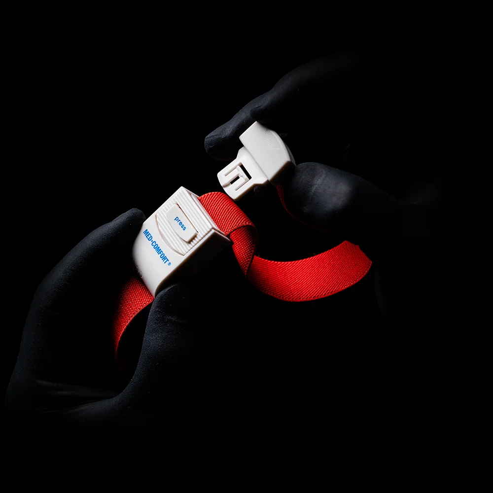 Mâini mănușate, care amintesc de un garou AMPri MED-COMFORT, apucă catarama unei centuri de siguranță roșu aprins pe un fundal umbrit. Pe cataramă se pot vedea cuvântul „Blue” și litera „B”.