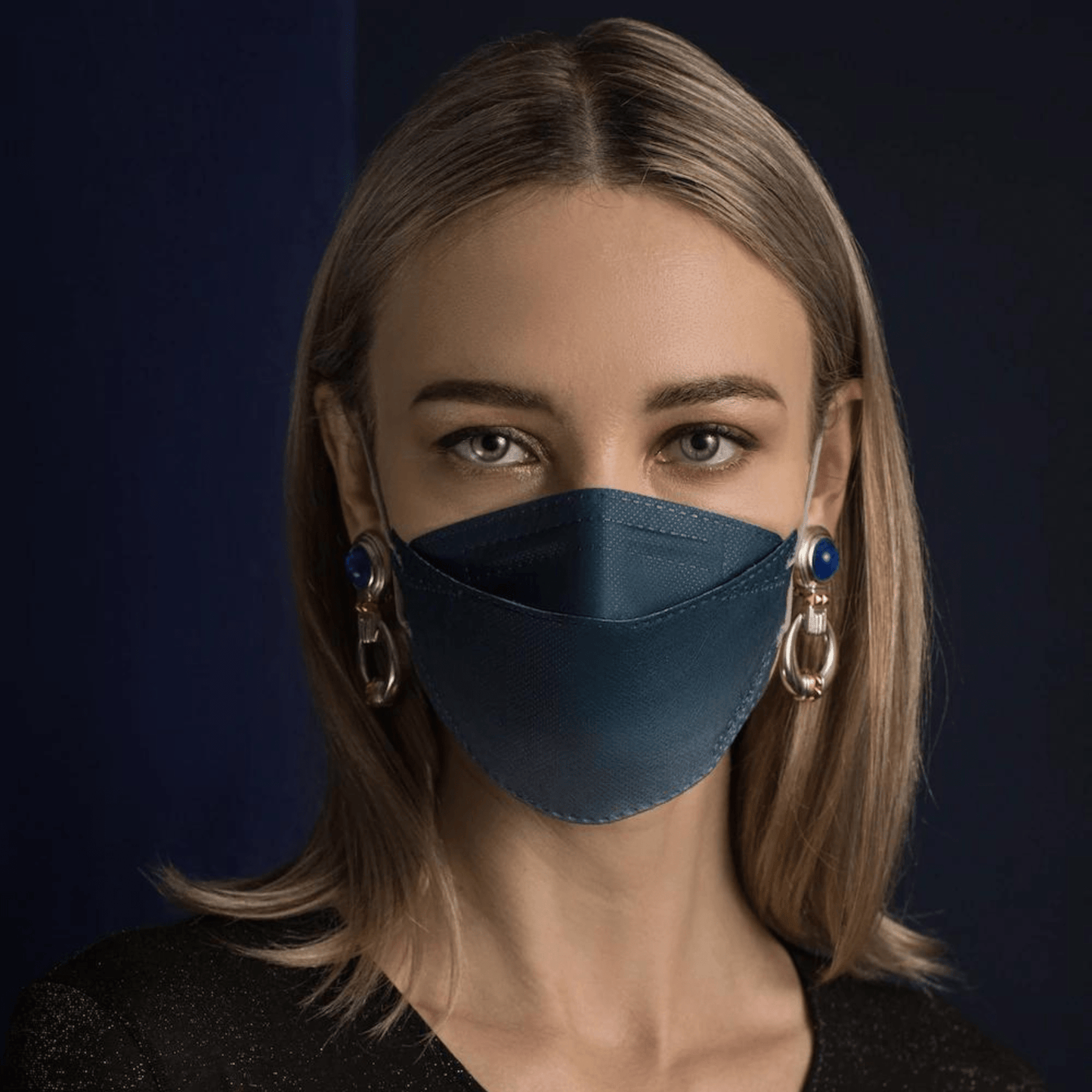 O persoană cu părul lung poartă o mască de față Altruan cu filtru de particule în albastru, care oferă protecție eficientă conform standardului FFP2. O combină cu cercei mari decorativi pe un fundal întunecat și poartă o cămașă neagră.
