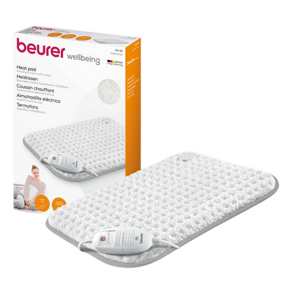 Lângă produs este ilustrat un Beurer HK 42 pernă electrică Super-Cosy de la Beurer GmbH cu marca „wellbeing“ pe ambalaj. Perna albă cu margine gri este echipată cu o telecomandă. Ambalajul arată de asemenea o imagine a pernei electrice în utilizare și evidențiază funcția de încălzire rapidă.