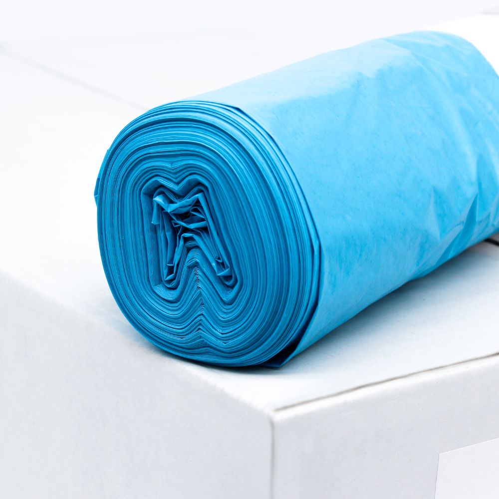 Un sul de saci de gunoi albaștri DEISS LDPE tip 60, 20772, 70 litri de la EMIL DEISS KG (GmbH + Co.) se află pe o cutie albă. Acești saci durabili, cunoscuți pentru rezistența lor la rupere, sunt rulați ordonat pentru a face vizibilă structura stratului materialului reciclat. Fundalul rămâne simplu și minimalist.