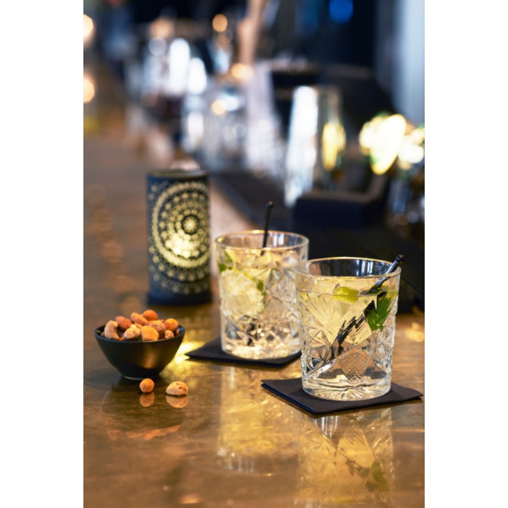 Zwei Gläser mit Eis, Limettenscheiben und Strohhalmen stehen auf quadratischen Untersetzern auf einer Bartheke. Daneben befindet sich eine kleine schwarze Schale mit gemischten Nüssen. Der Duni Kerzenhalter Billy von Duni GmbH, aus schlichtem Metall 140 x 75 mm mit modernem Design, leuchtet sanft im Hintergrund und schafft eine warme Atmosphäre.