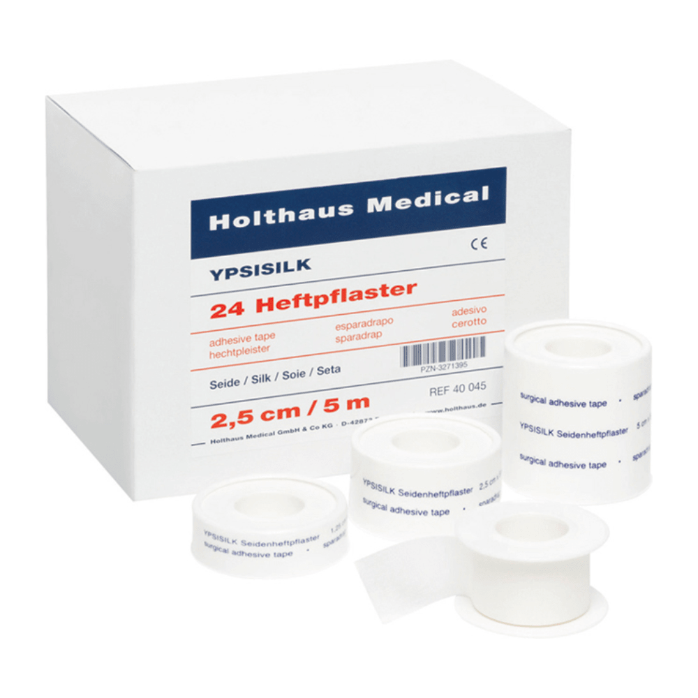 O cutie cu inscripția „Holthaus Medical GmbH & Co. KG“ cu „Holthaus YPSISILK bandă adezivă – 2,5 cm x 5 m | Pachet (5 m)“ și mai multe role de bandă adezivă albă în diferite dimensiuni în fața ei. Textul de pe cutie și benzi conține detalii despre produs în mai multe limbi.