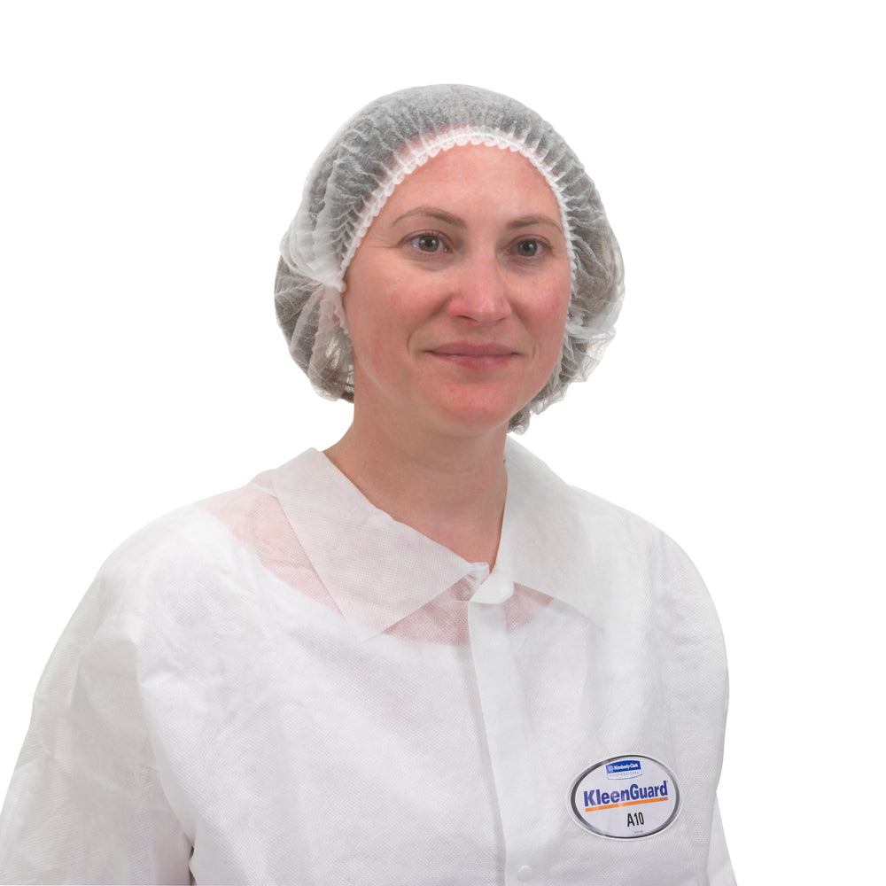 Eine Person trägt einen weißen Einweglaborkittel und eine KleenGuard® A10 Leichte Haube mit Gummizug (von Kimberly-Clark GmbH) mit einem "KleenGuard"-Namensschild vor einem schlichten weißen Hintergrund.