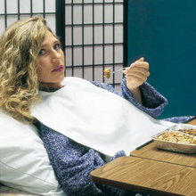 Eine Person mit lockigem Haar liegt im Bett an einem Kissen gelehnt, trägt einen blauen Pullover und einen Servocare Kleiderschutz für Erwachsene von Servoprax GmbH. Sie isst mit einer Gabel Essen aus einem Folienbehälter. Vor ihnen steht ein Holztisch und im Hintergrund ein Shoji-Paravent.