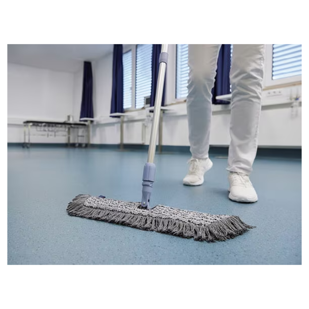 O persoană în pantofi și pantaloni albi șterge un podea albastră cu un mop Vileda GmbH Professional Swep Duo r-MicroCombi - 50 cm, într-o cameră cu jaluzele închise și perdele albastre. În apropiere se află o masă pliantă și un calorifer sub ferestre.
