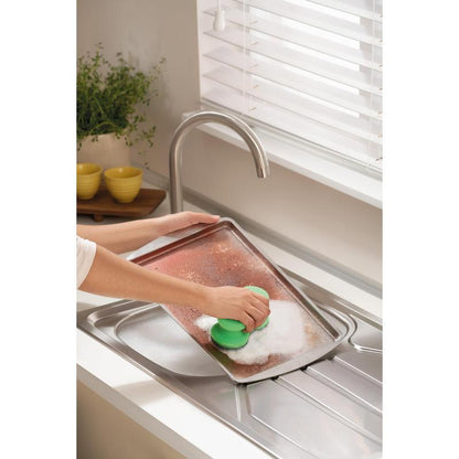 Cineva freacă o tavă mare și murdară cu buretele de curățare Scotch-Brite® Classic de la 3M Deutschland GmbH sub un robinet într-o bucătărie luminoasă cu jaluzele albe, o plantă și boluri galbene stivuite în fundal.